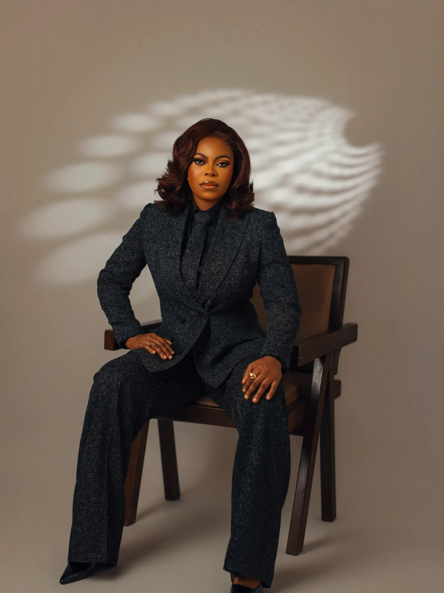 Woman in a charcoal tweed suit seated on a wooden chair, leaf shadows on the wall behind